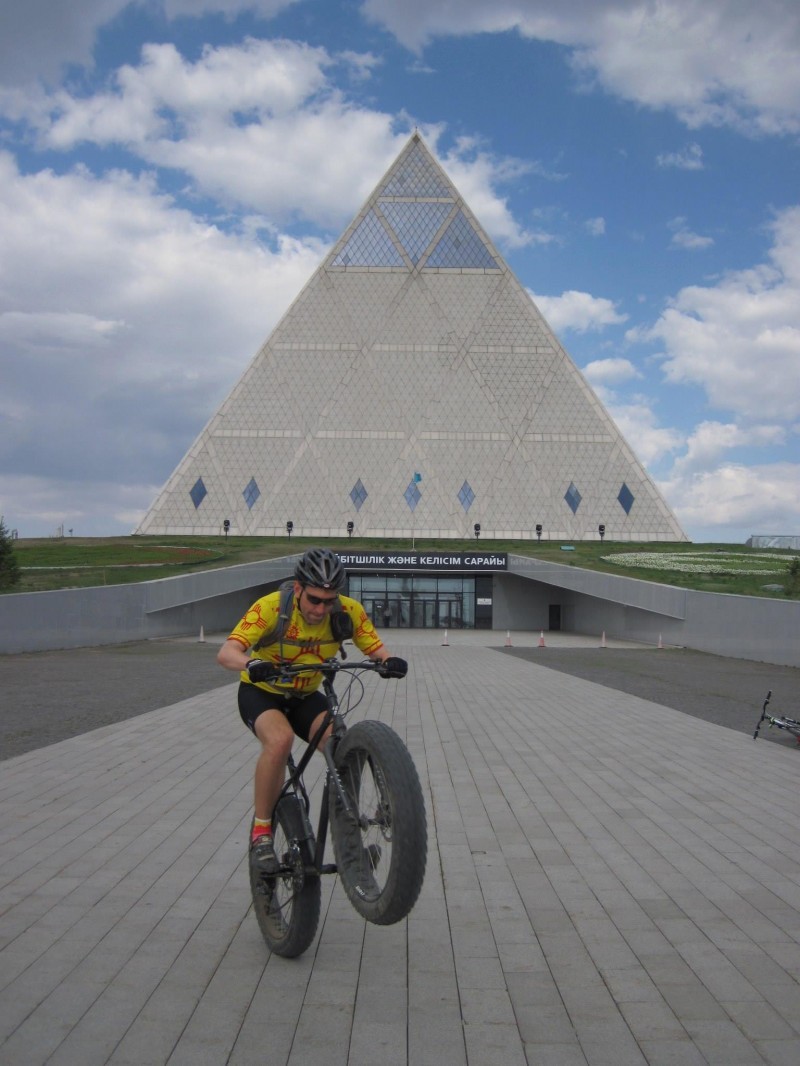 Front view of a cyclist, riding a wheelie on a black Surly Moonlander fat bike, up a ramp in front of a pyramid building