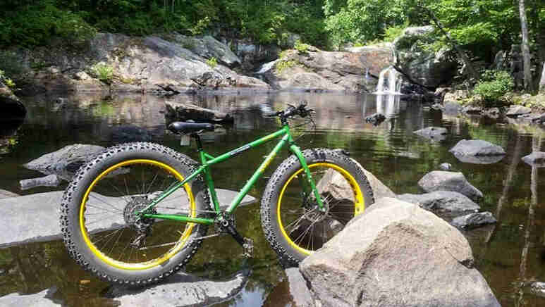 Right side view of a green Surly Moonlander fat bike, parked on large rocks in a pond with a waterfall in the background