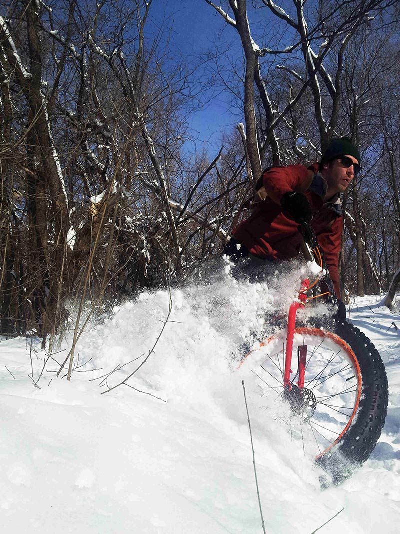 Front, right side view of cyclist, riding a red Surly Moonlander fat bike, kicking up snow on a snowy trail in the woods