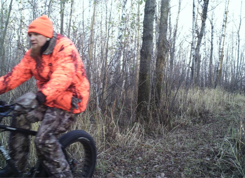 Left side view of a cyclist in blaze orange outerwear, riding a Surly Moonlander fat bike through the bare woods