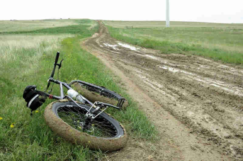 Rear view of a Surly Pugsley fat bike, laying on it's left side in grass field next to a muddy road
