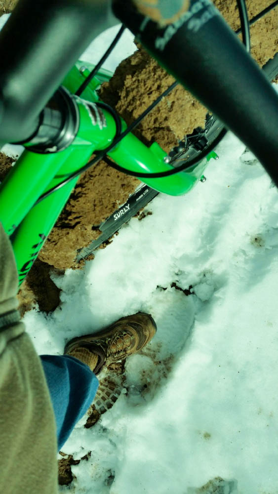 Cropped, downward view of a green Surly fat bike with a muddy front tire, parked in snow next to a person's shoe