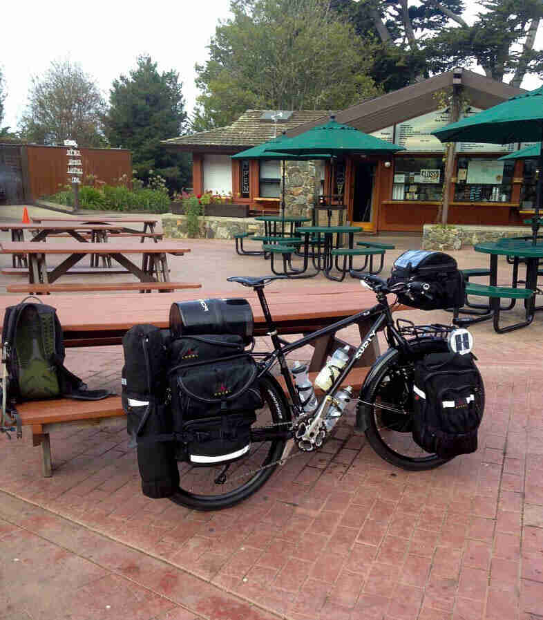 Right side view of a black Surly Troll bike, loaded with gear, parked against a picnic table on an outdoor food court