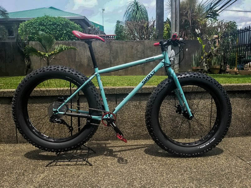 Right side view of a mint Surly Wednesday fat bike, parked on a sidewalk next to a stone ledge with a grass yard on top