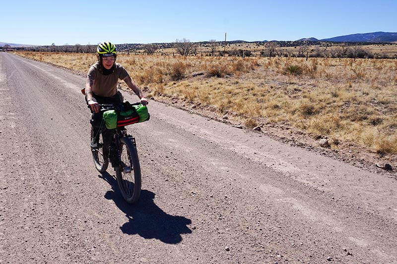 Front view of Elena riding a gear packed Surly bike down a gravel road in a brushy desert with hill in the distance