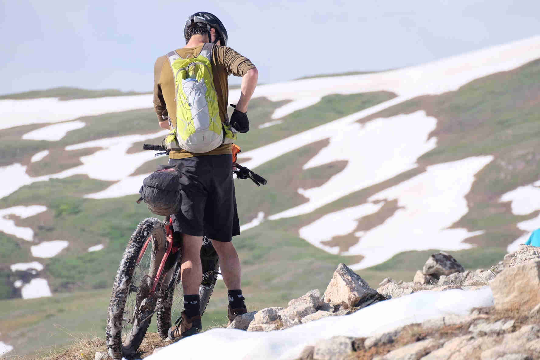 Rear view of a cyclist standing with a bike while adjusting a backpack strap, on a rocky hill in the mountains