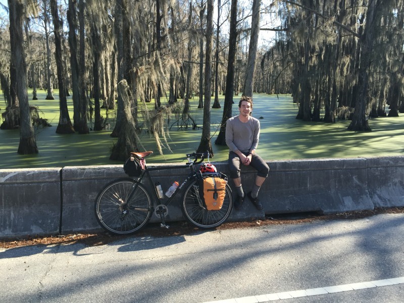 Right side view of a black Surly bike, parked along a barrier with cyclist sitting on top, with a swamp forest behind