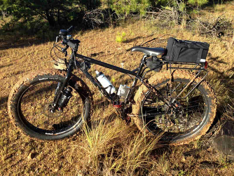 Left side view of a muddy, black Surly Pugsley fat bike with a rear pack, parked on a grass field