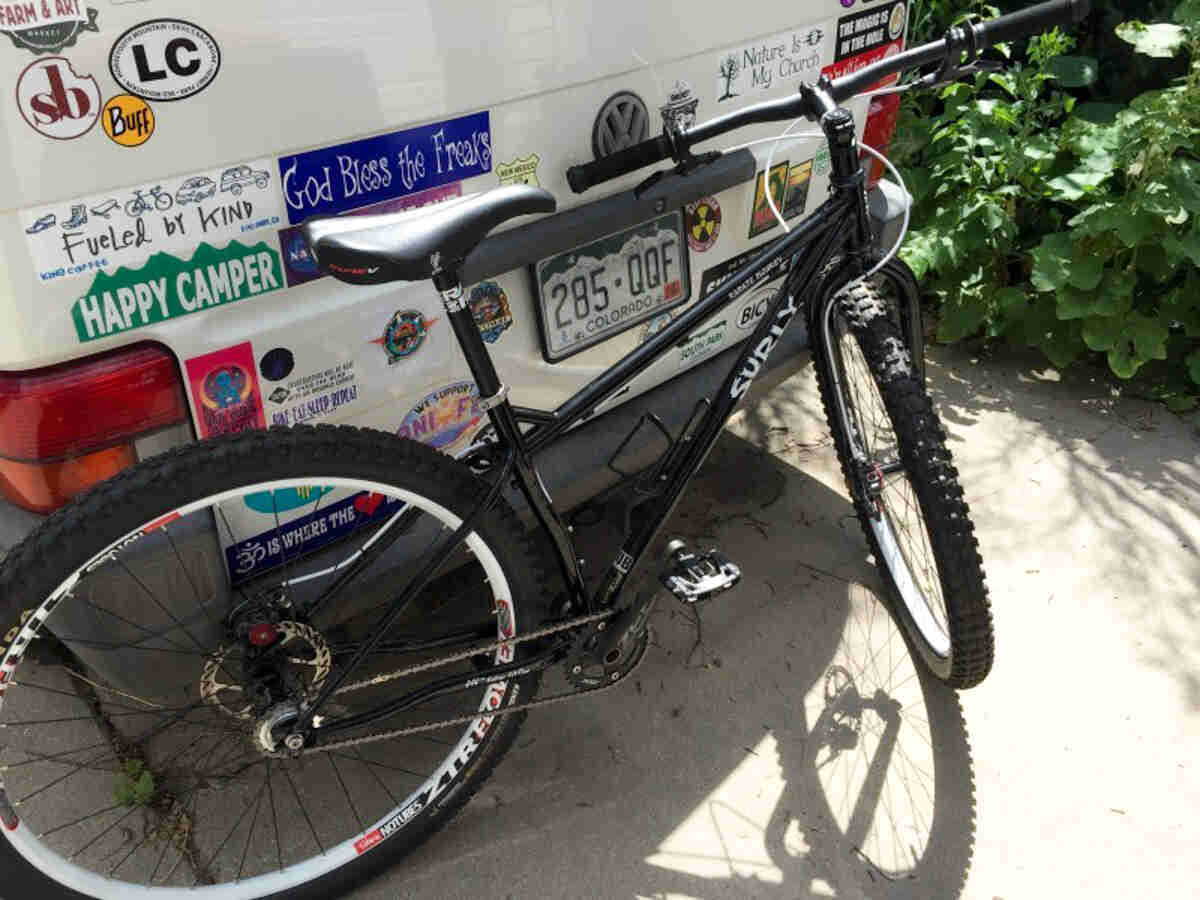 Downward, right side view of a black Surly Karate Monkey bike, parked against the rear of a VW van with stickers on it
