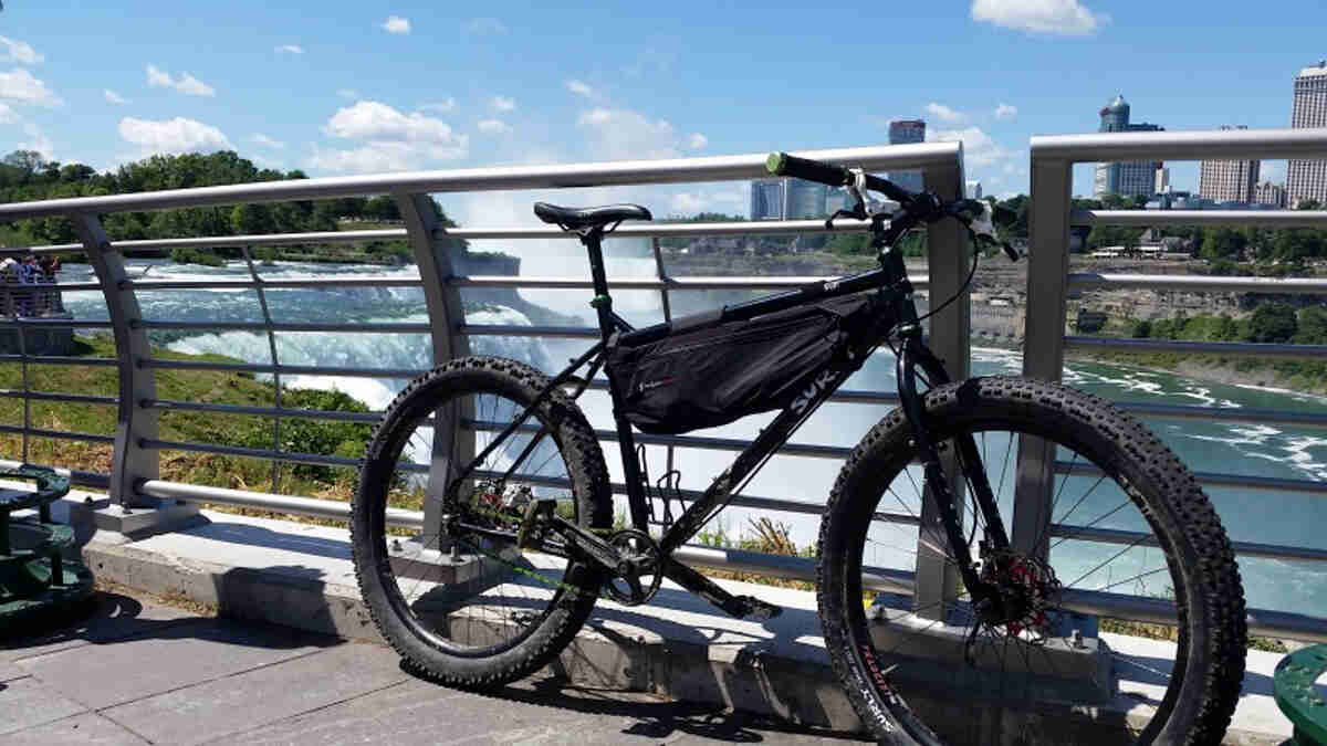 Right side view of a black Surly Troll bike, parked against a railing, with a large waterfall and city in the background