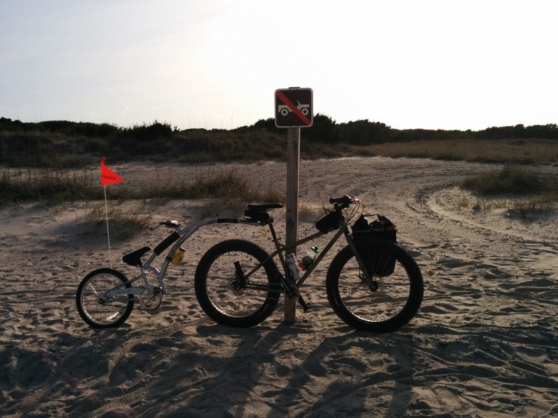 Right side view of a Surly fat bike, with a child's tag along trailer, leaning against a signpost in a sandy field