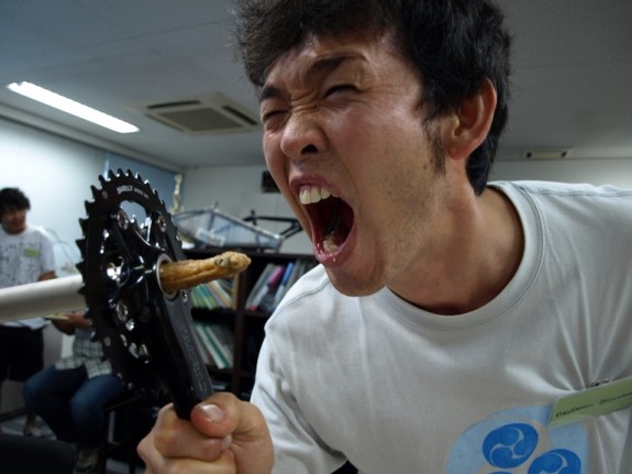 Headshot of a person with their mouth wide open, holding a Surly O.D bike crank, with a chicken wing sticking out of it