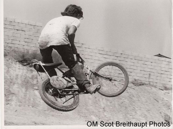 Right side view of a cyclist in the air on a banana seat bike, with a wall on a hilltop, in background - black & white