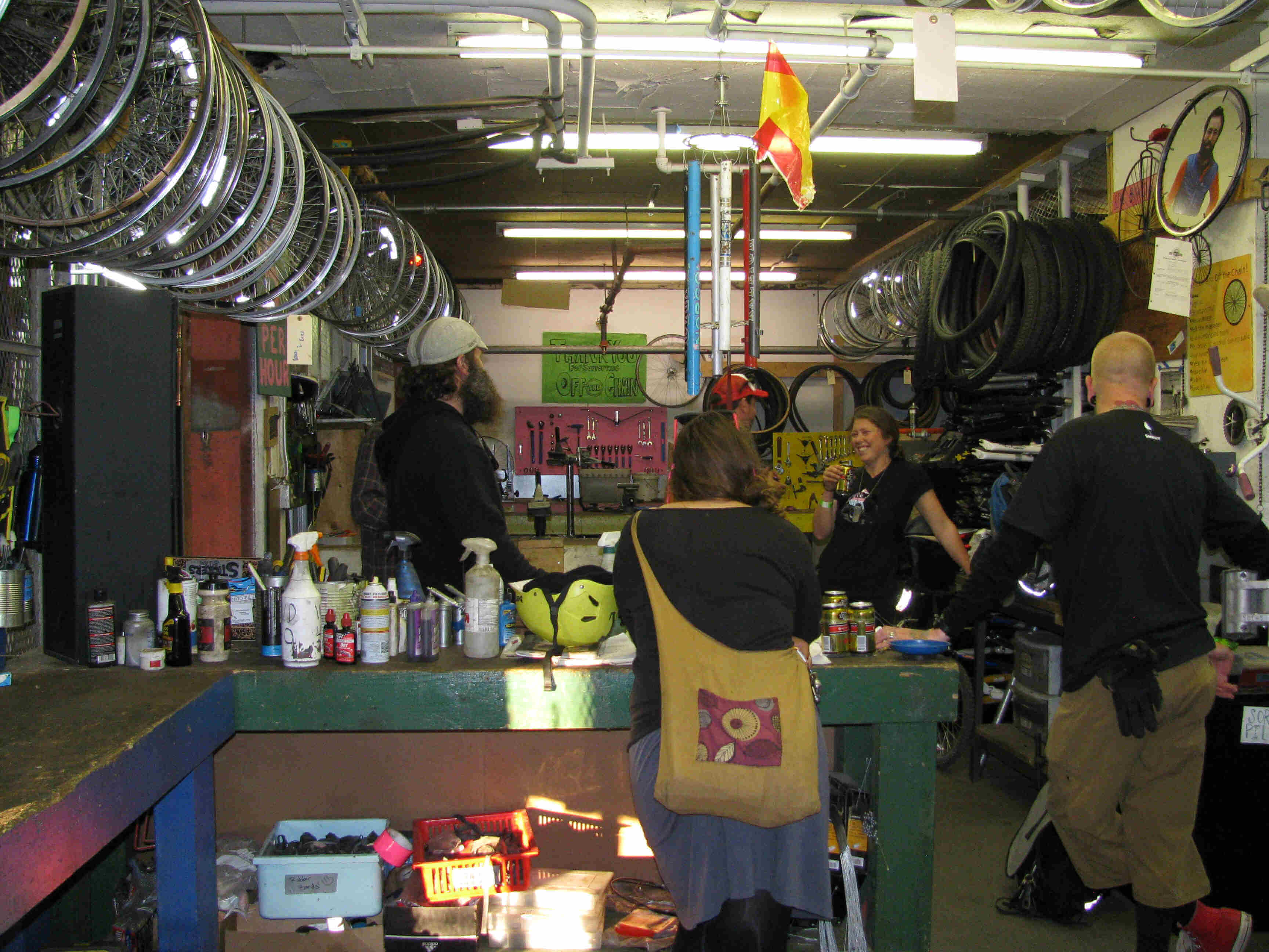 Rear view of 2 people standing near a workbench, talking to other people, in a bike shop