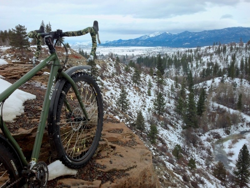 Rear, right side view of an green Surly Ogre bike, on a cliff edge, overlooking a snowy valley of trees in the mountains