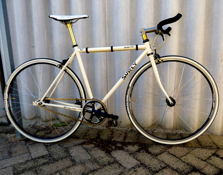 Right side view of a white Surly Steamroller bike, leaning against a white, steel wall