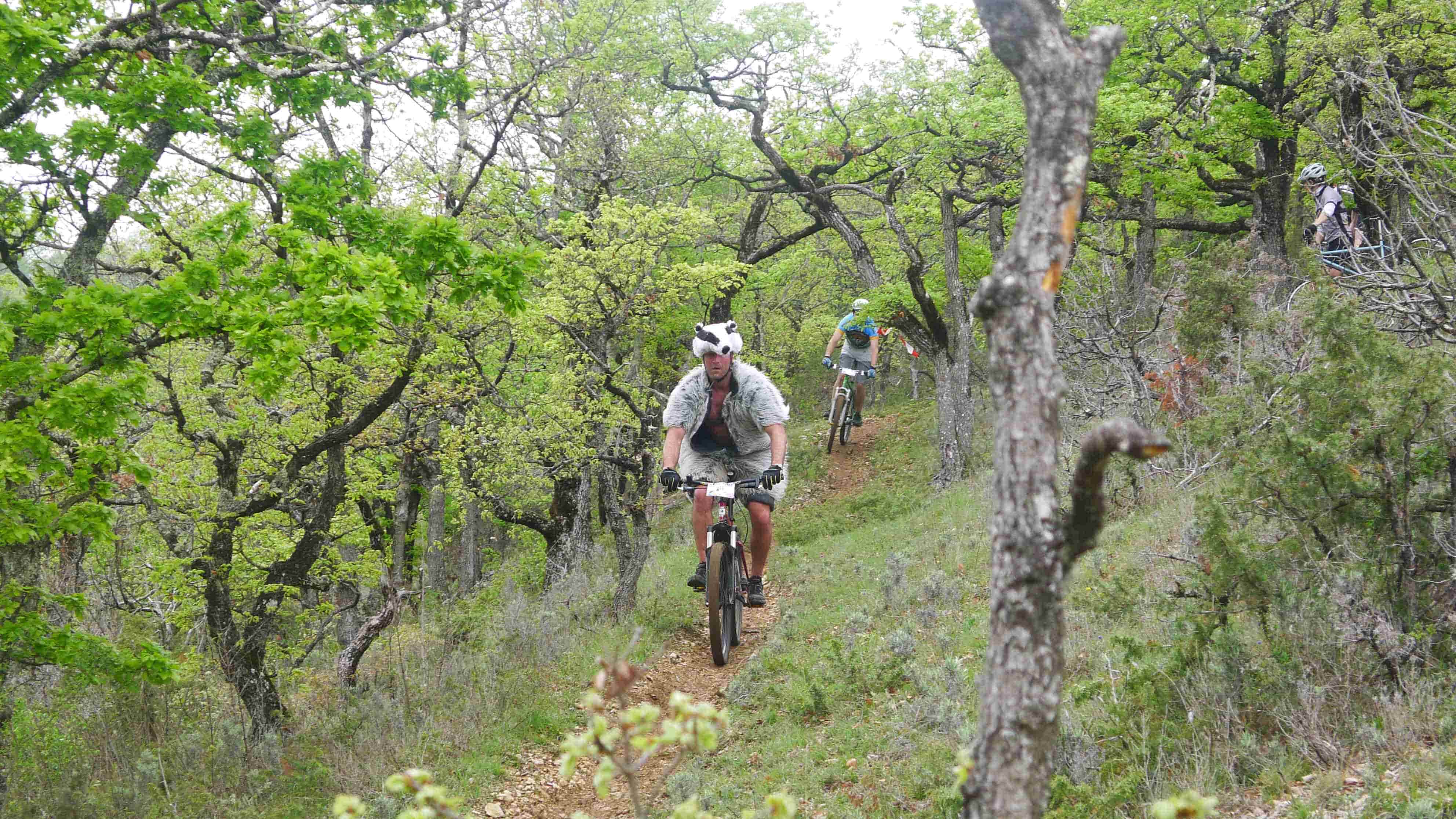 Front view of a cyclist, riding their bike on a trail with another rider behind them, in the grassy woods