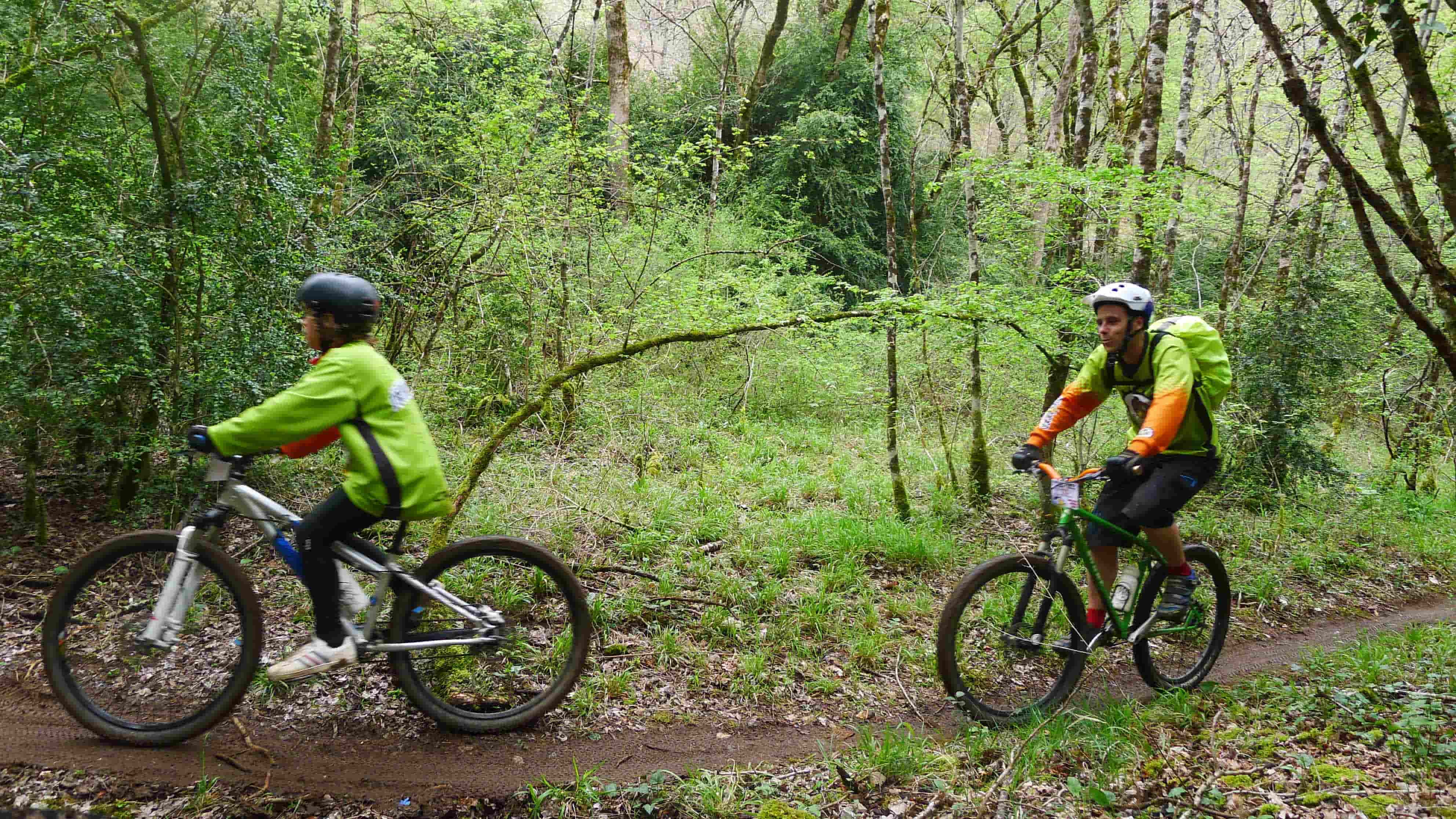 Left side of 2 young cyclists, riding on their bike in single file, on a dirt trail in the woods