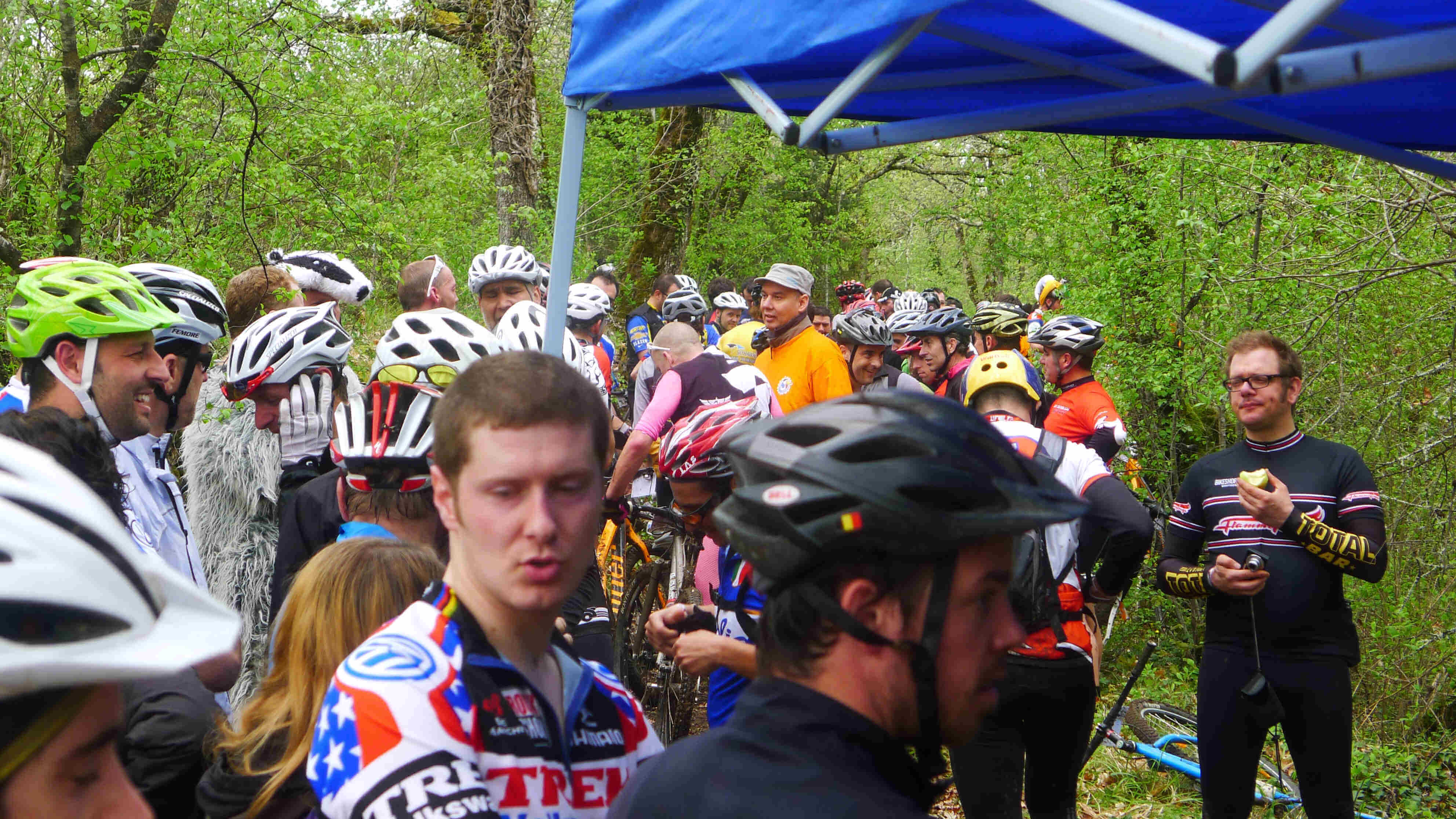 A chest up view of a group of cyclists, standing near a blue canopy, in the woods
