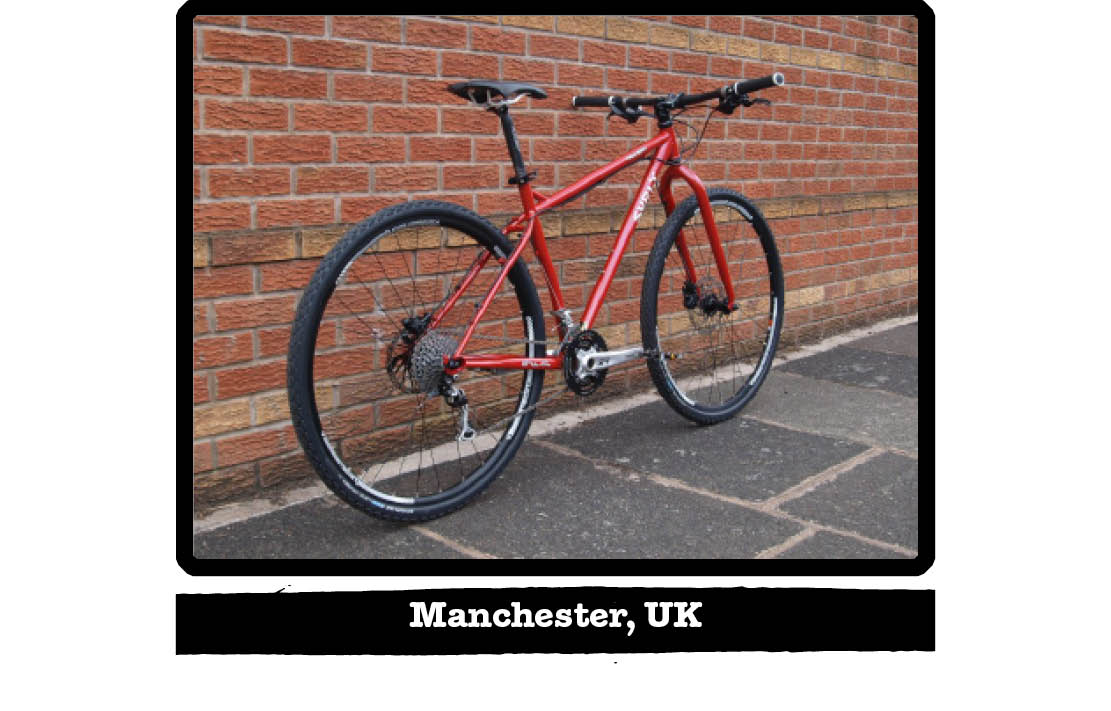 Rear right side view of a red Surly bike on a sidewalk, leaning on a red brick wall - Manchester, UK tag below image