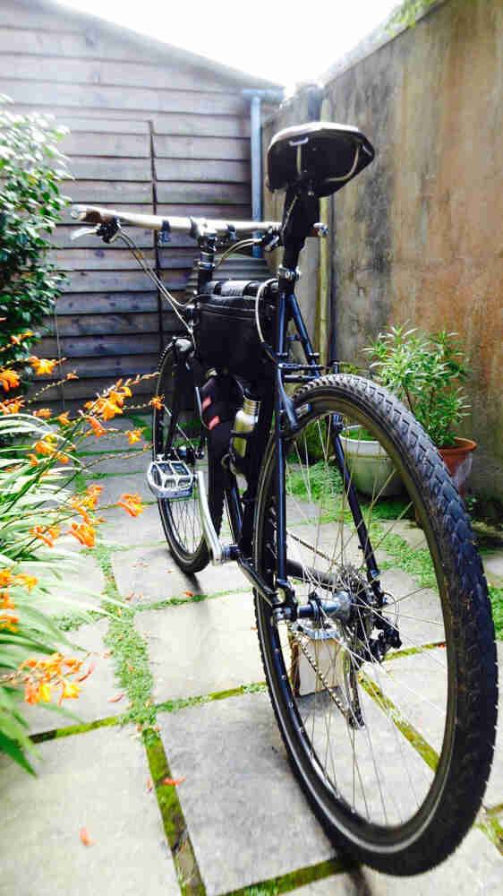 Rear view of a black bike with frame pack, parked on a block walkway, in front of a shed