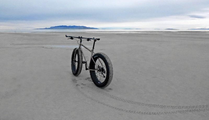 Rear, left side view of a tan Surly fat bike, standing in a flat sand area, facing a body of water on a cloudy day