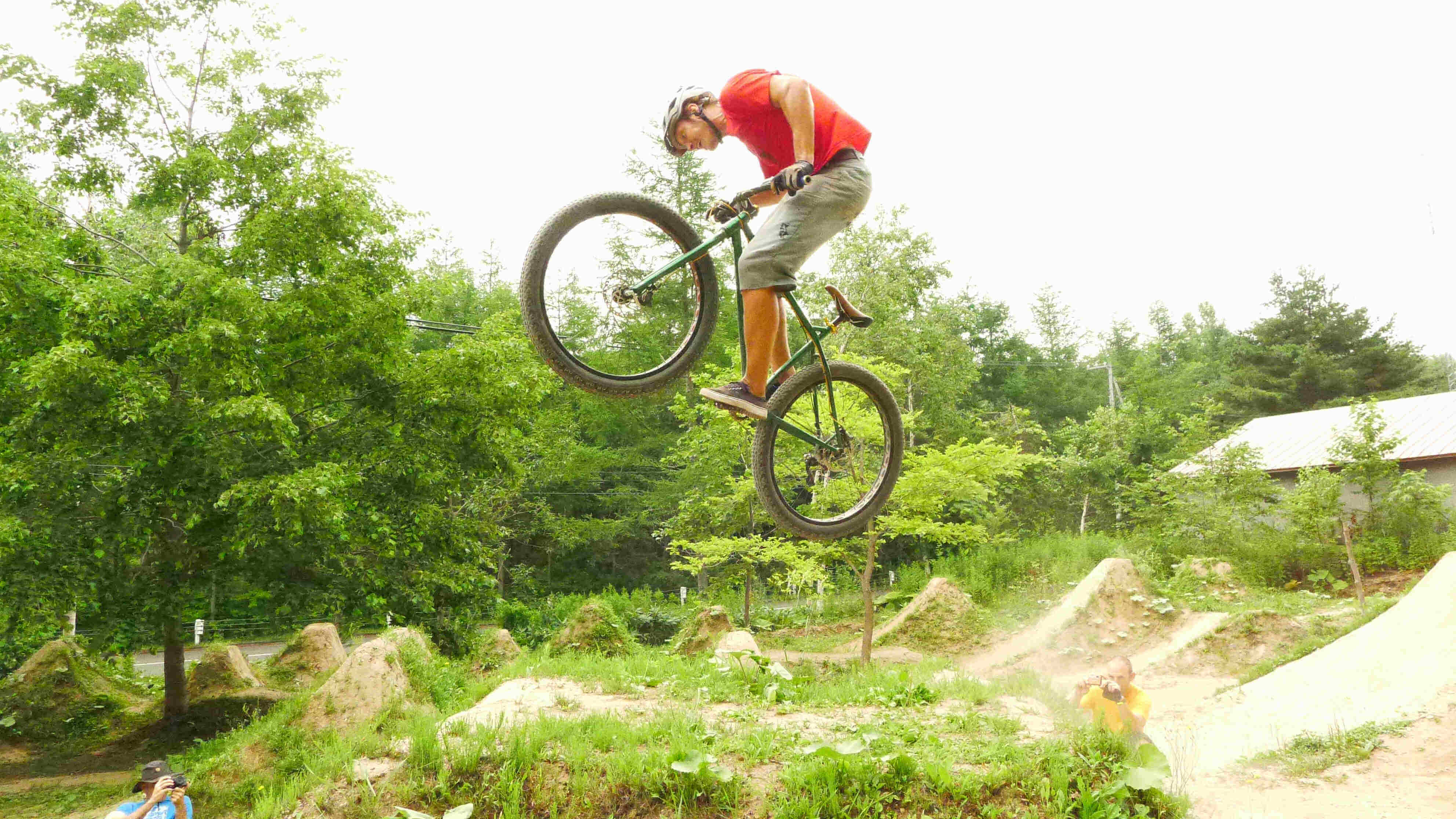 Left side view of a cyclist, riding a green Surly Krampus bike, flying off of a dirt ramp on a BMX track in the woods