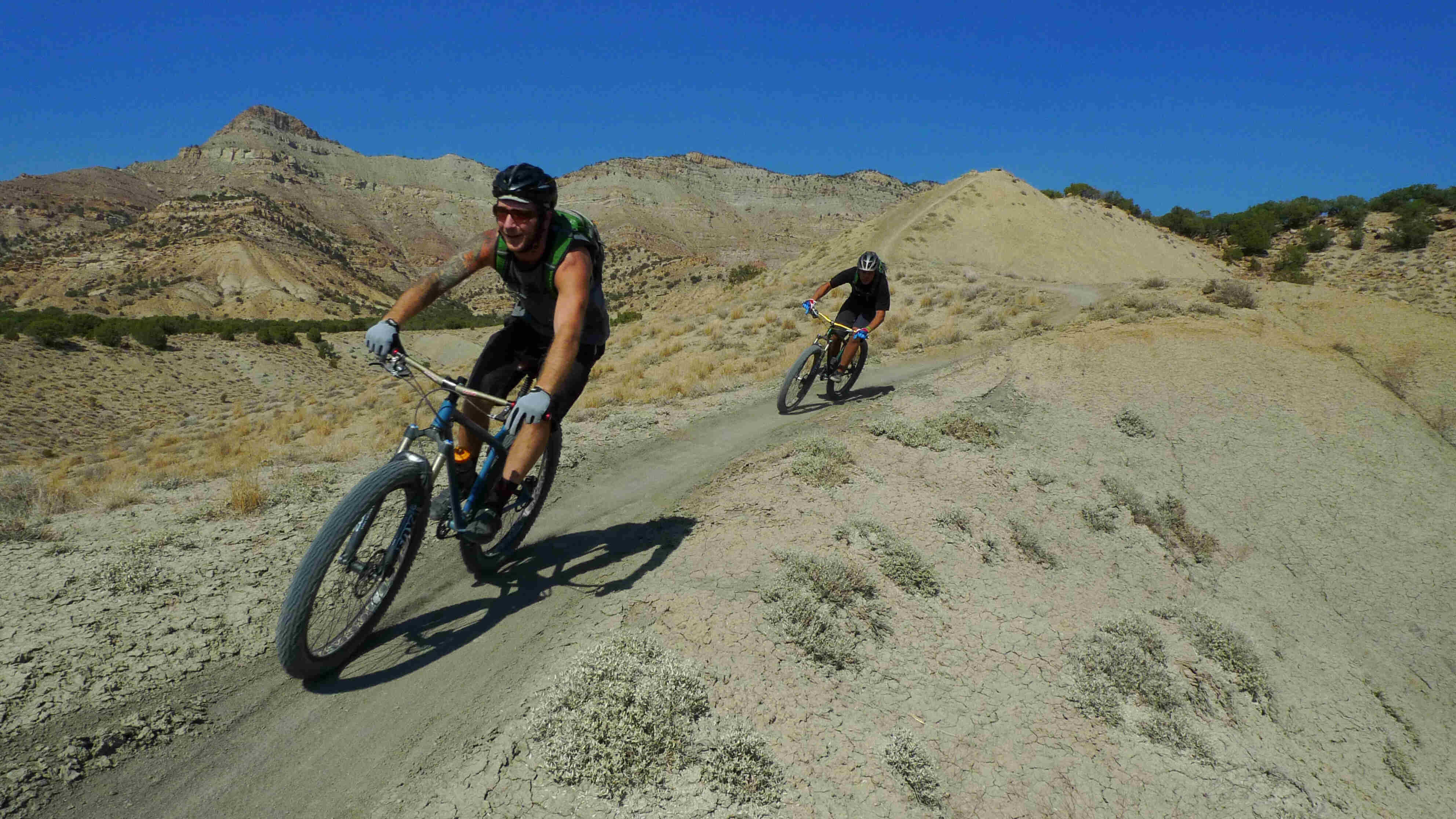 Front, left side view of 2 cyclists on Surly bikes, riding single file, down a trail on sandy, rock hills