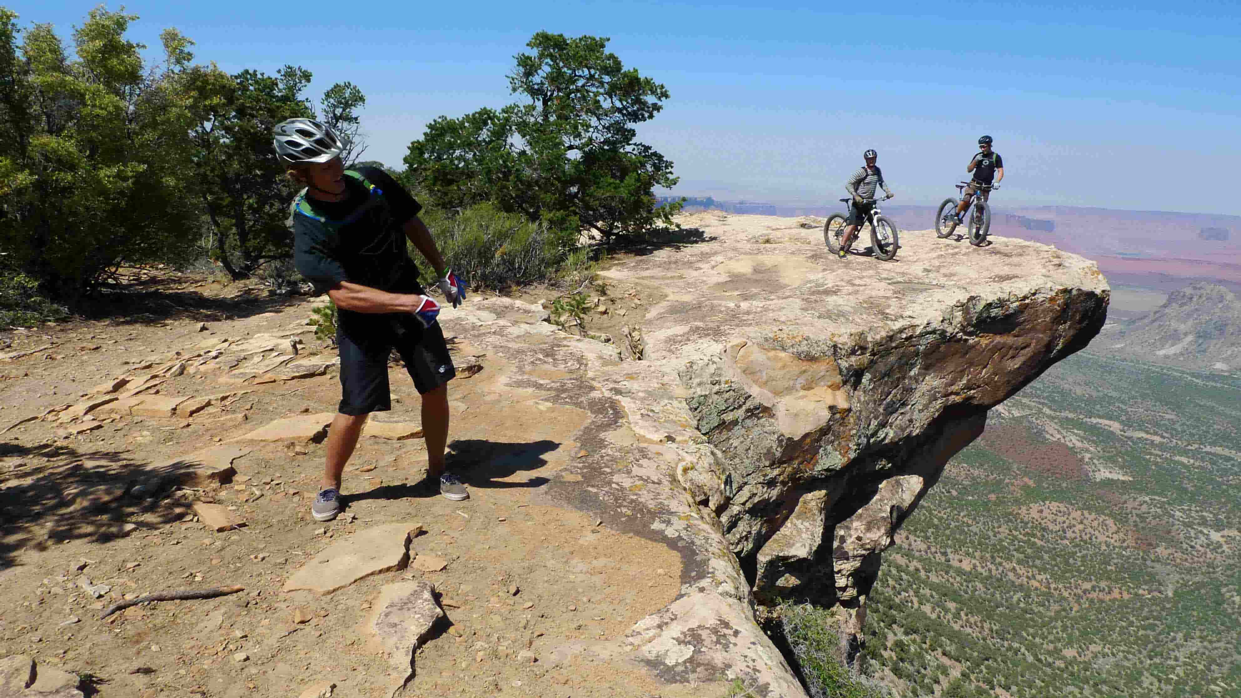Front view of a cyclist standing near the edge of a high, rock cliff, with 2 cyclists and their Surly bike, behind them