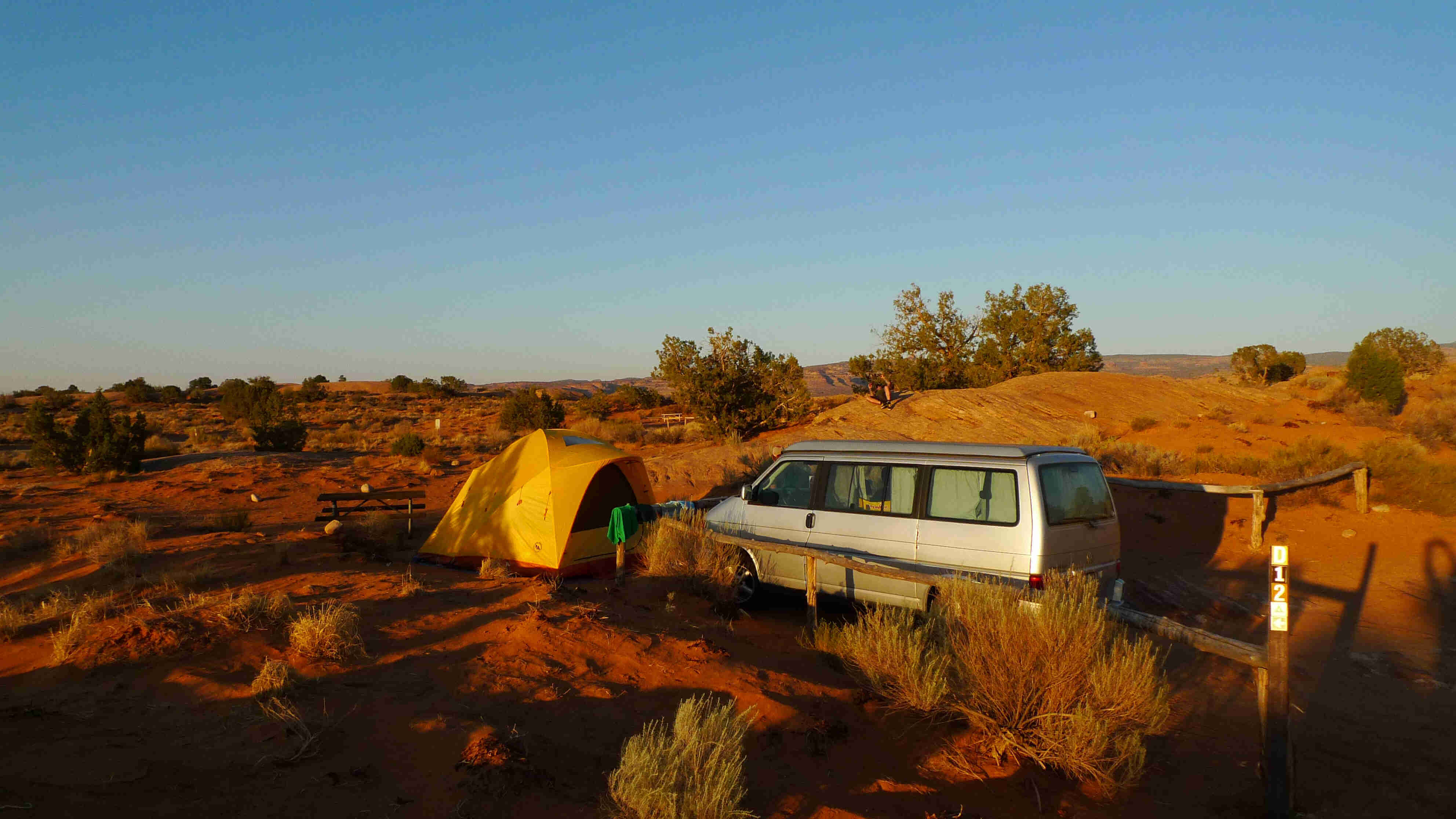 Left side view of a silver van, with a yellow tent set up near the front bumper, in a brushy desert field