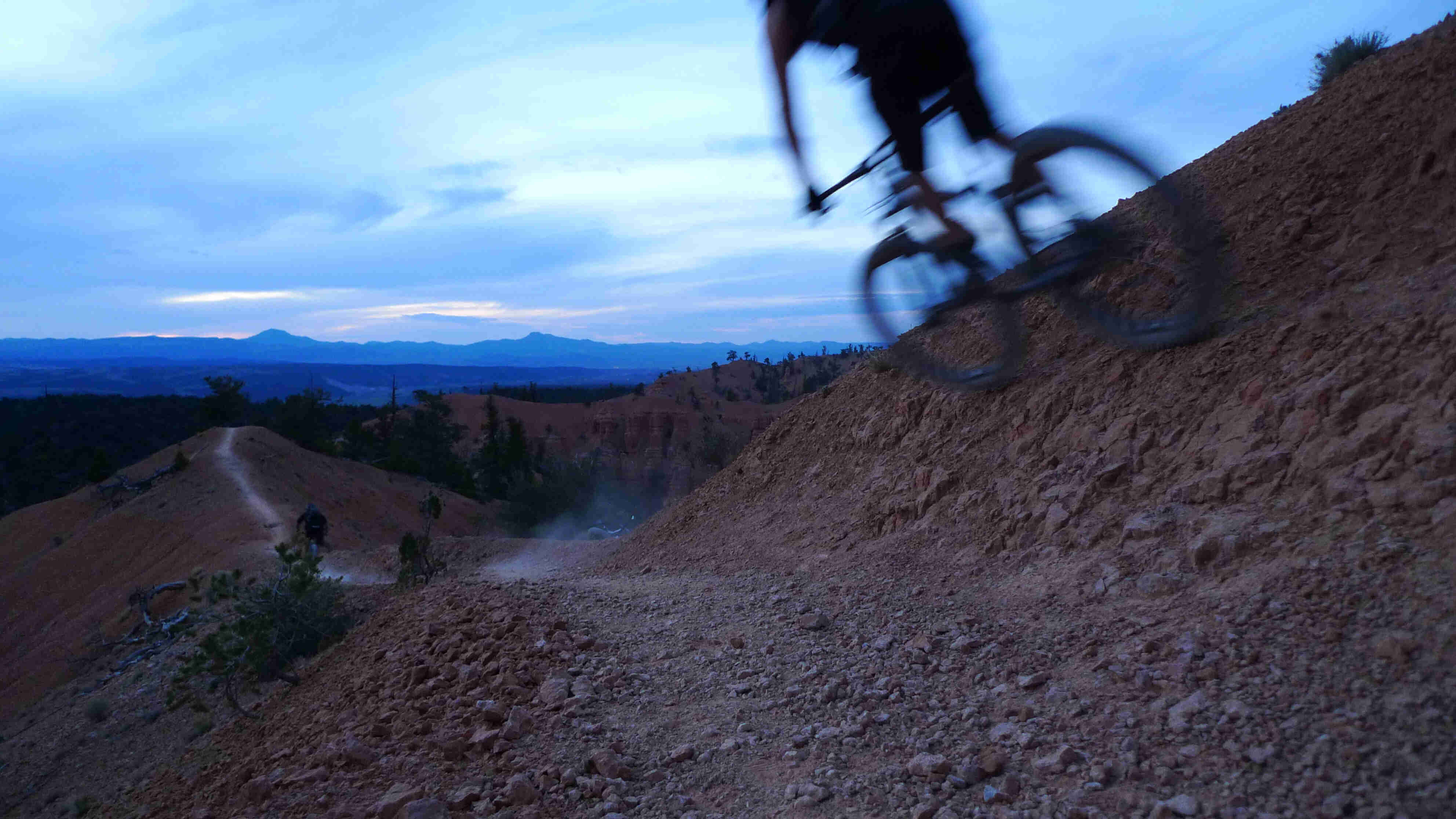 Rear, left side, blurry view of a cyclist, riding across a rocky hill, on a trail in the bare mountains