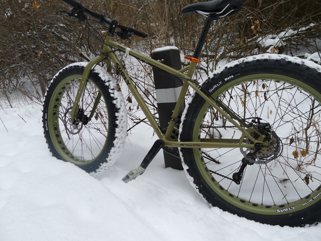 Left view of an olive green Surly Pugsley fat bike, parked on snow, leaning against a wood post