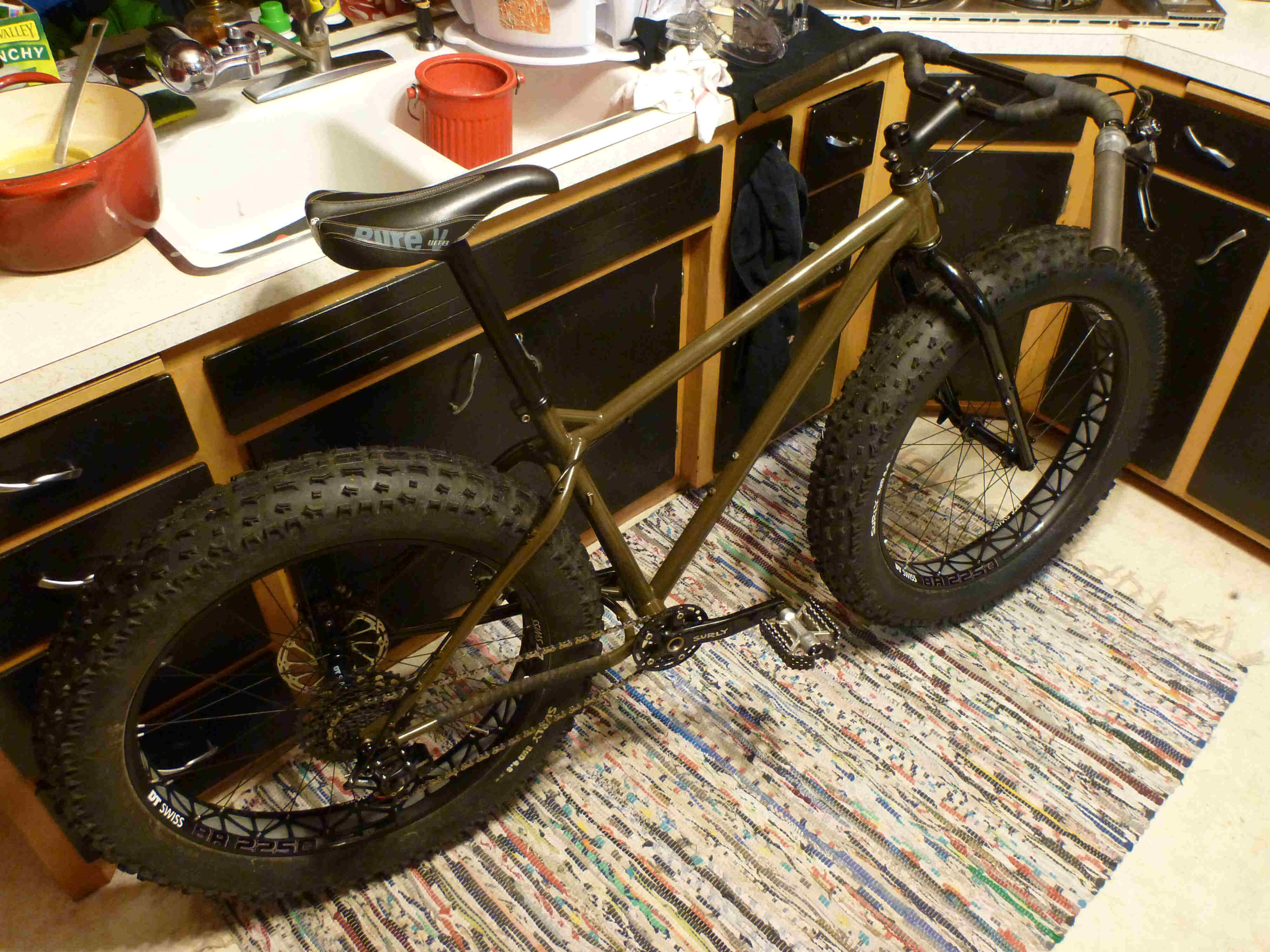 Downward, right side view of an olive drab Surly Ice Cream Truck fat bike, leaning against cabinets in a kitchen