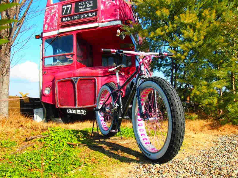 Front, right side view of a Surly fat bike with white wall tires, parked in front of a red, double decker bus and a tree