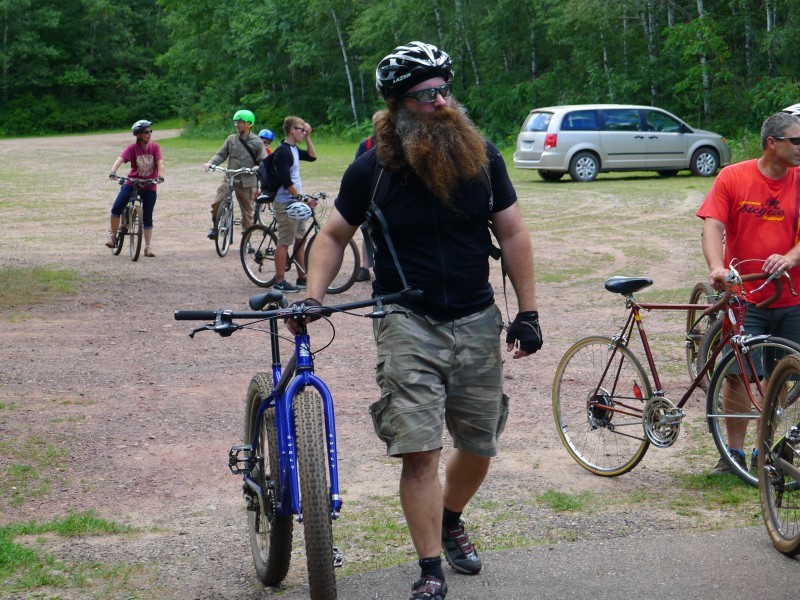 Front view of a cyclist with a thick beard, walking with a bike on a gravel lot, with other cyclists in the background