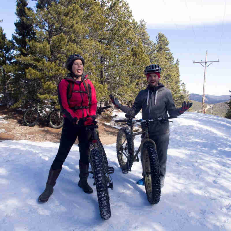 Front view of 2 cyclists, posing with their Surly fat bikes, standing on snowy ground, with pine trees behind them