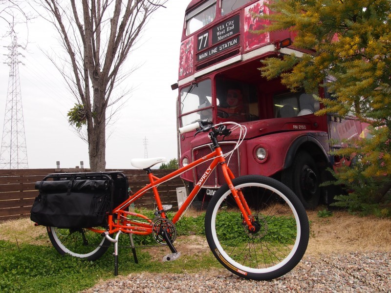 Right side view of a modified, red Surly Troll with a rear rack, in front of a tree, wood fence, and the front of a bus