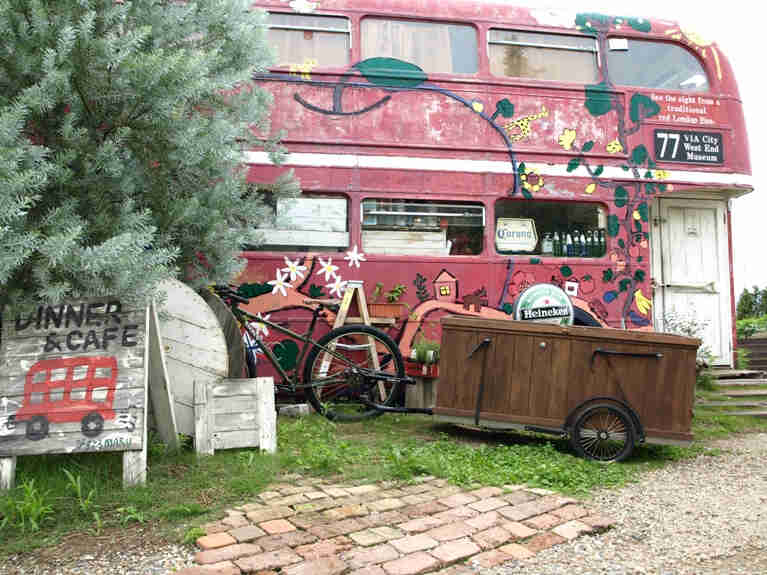 Left side view of a Surly bike with a trailer, parked on the side of a double decker bus that was converted into a diner