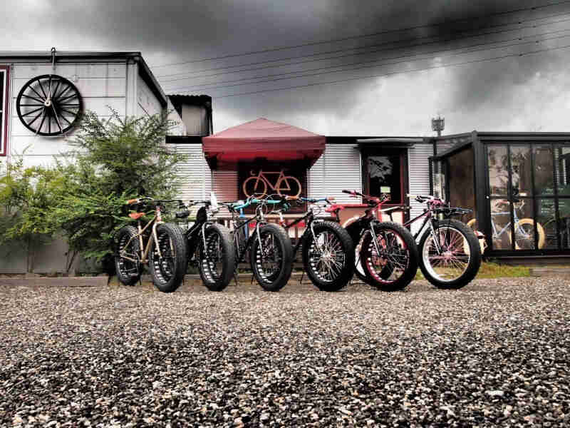 Front, right side view of a row of Surly fat bikes, sided by side on a gravel parking lot, outside of a bike shop