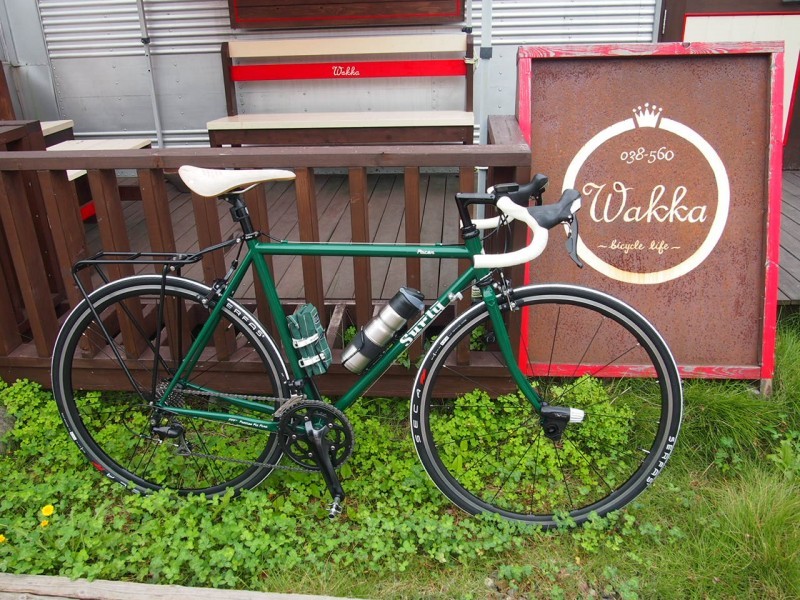 Right side view of a green Surly Pacer bike, parked on grass, outside of a wood deck patio with a steel building behind