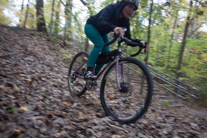 Front, right side view of a cyclist, on a purple Surly bike, riding down a leaf covered hill in the woods