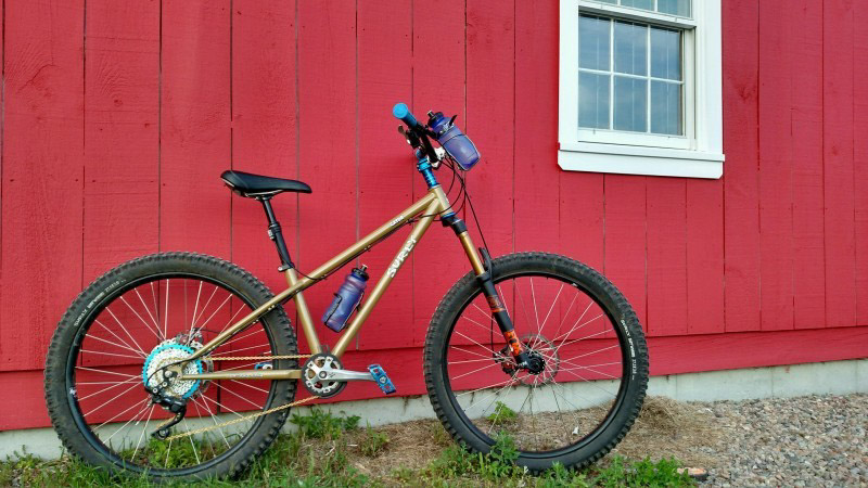 Right side view of a Surly bike, gold, parked next to the wall of a red barn