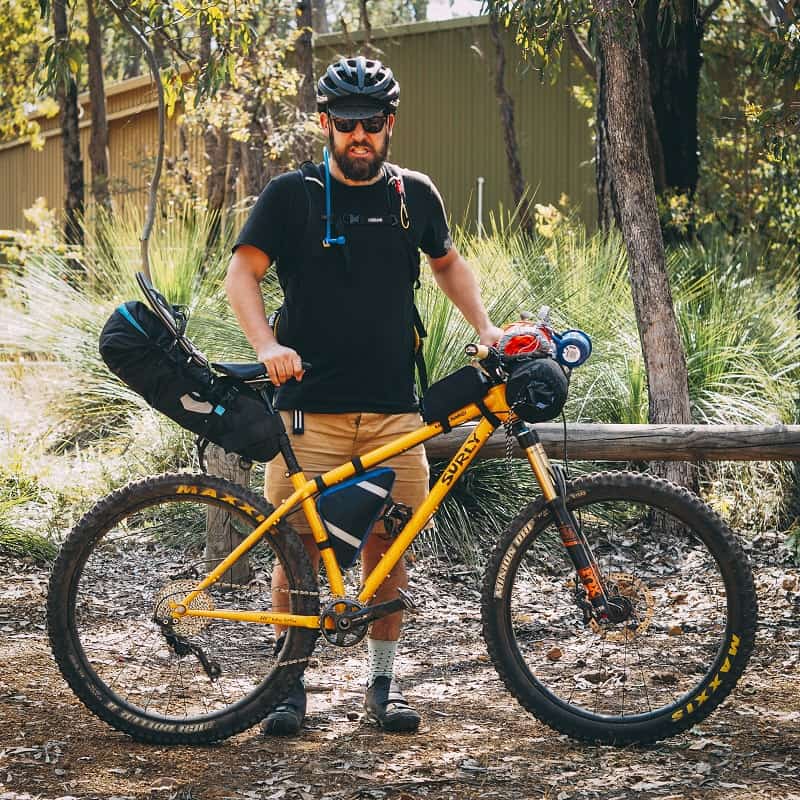 Right side view of a yellow Surly Karate Monkey bike with cyclist behind, and tall grass and trees in background