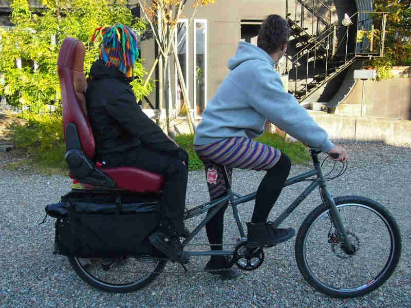 Right side view of a Surly Big Dummy bike, with cyclist on front, and a person sitting on a car seat on the rear rack