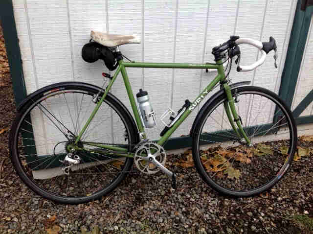Right side view of a green Surly Cross Check bike, parked on gravel, against the side of a gray woodshed