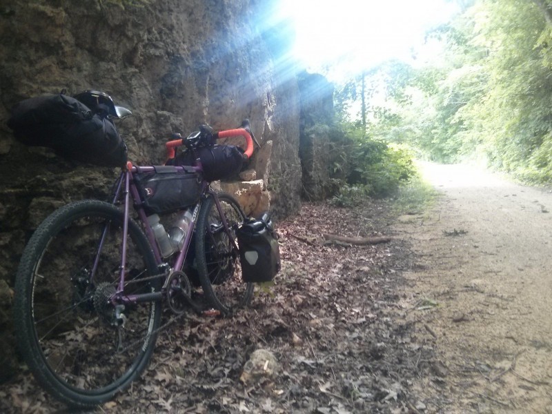 Rear, right side view of a purple Surly bike with gear, leaning on a cliff wall base, next to a gravel road in the woods
