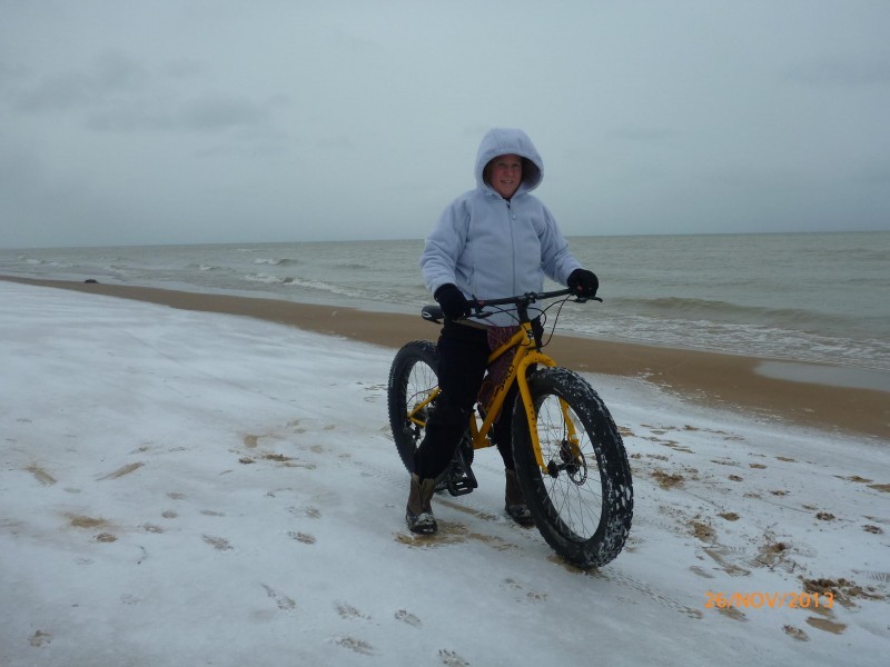 Front, right side view of a yellow Surly Pugsley fat bike with cyclist standing over, on a snowy shore with ocean behind