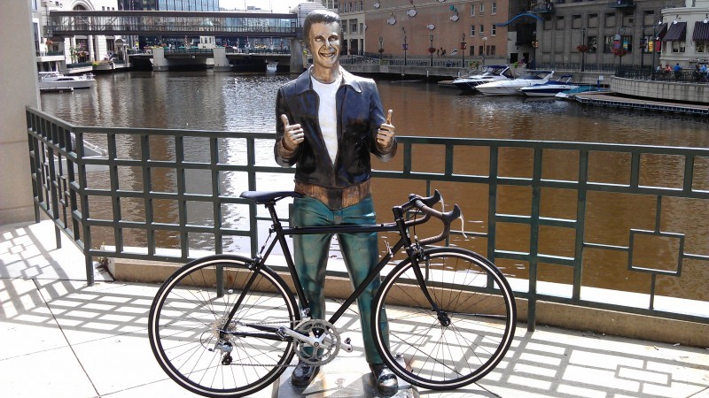 Right side view of a black Surly Pacer bike, against a statue of The Fonz, in front of a rail with a canal behind