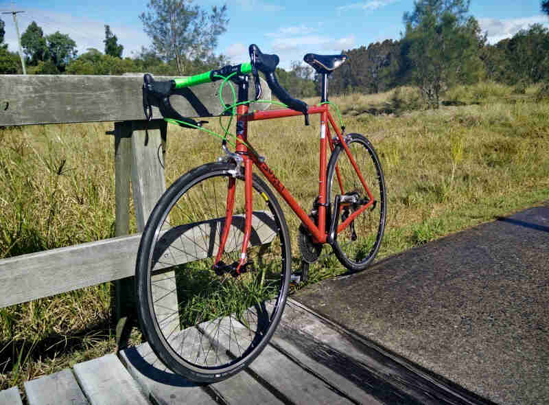 Left side view of a red Surly Pacer bike, parked against a handrail on a trailside, with field of grass and trees behind