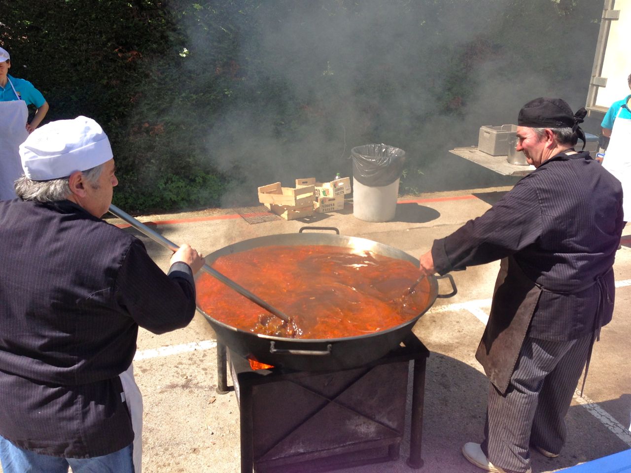 Side view of 2 people, standing and facing towards each other, stirring a large pan of food that's between them